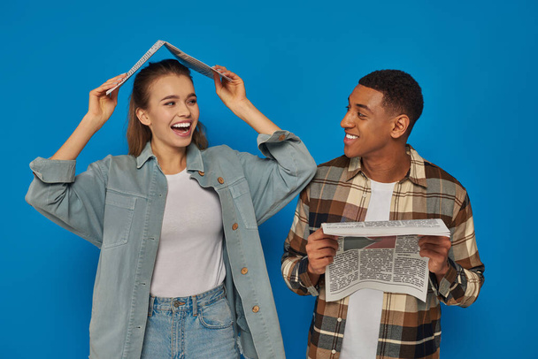 happy woman covering head with newspaper near smiling african american man on blue backdrop - Foto, immagini