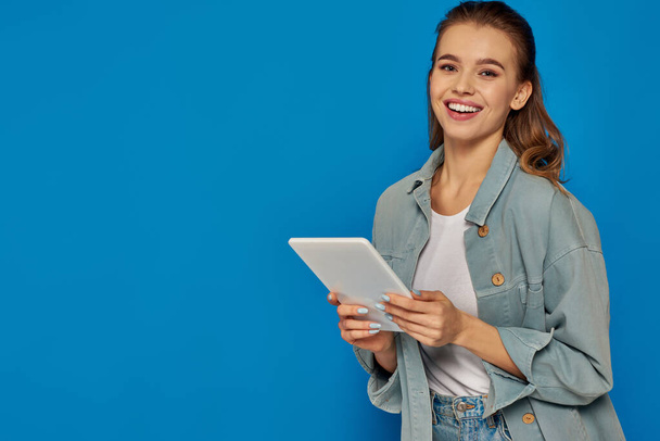gelukkig jong vrouw met digitale tablet en glimlachen op camera op blauwe achtergrond, social media - Foto, afbeelding