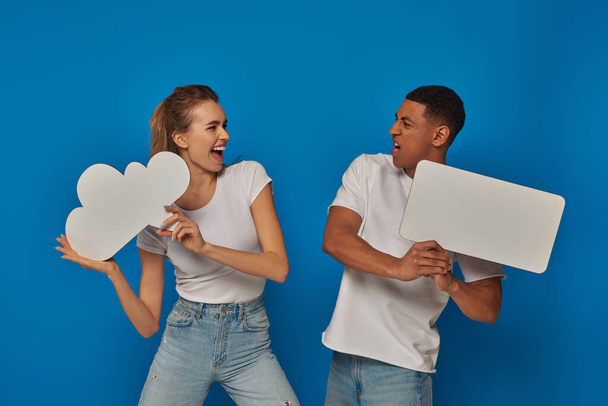 эмоциональная межрасовая пара держит пустые плакаты на синем фоне, мысле- и речевые пузыри - Фото, изображение