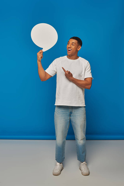 hombre afroamericano emocionado en vaqueros apuntando a la burbuja del habla en el fondo azul, longitud completa - Foto, Imagen