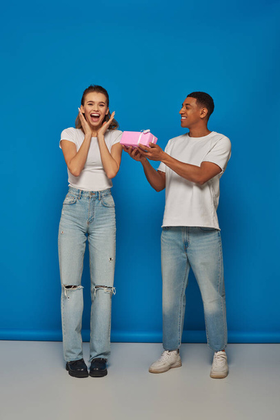 happy african american man presenting wrapped gift to excited girlfriend on blue backdrop - Photo, Image