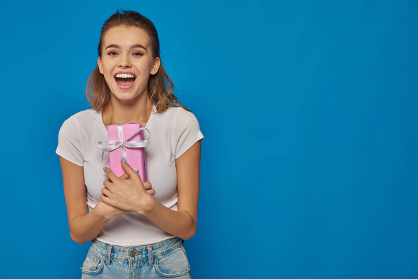 excited young woman holding gift box and looking at camera on blue background, festive occasions - Photo, Image