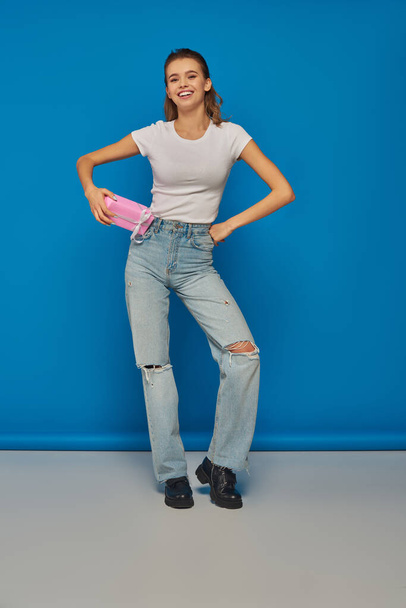 happy young woman in jeans holding gift and looking at camera on blue background, festive occasions - Photo, Image