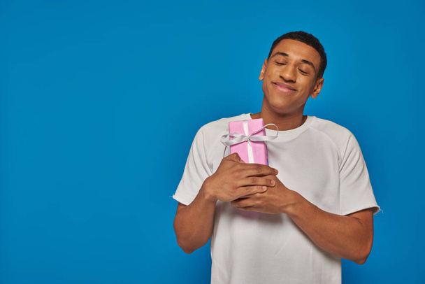 vreugdevolle Afro-Amerikaanse man glimlachend en met ingepakt geschenk op blauwe achtergrond, feestelijke gelegenheden - Foto, afbeelding