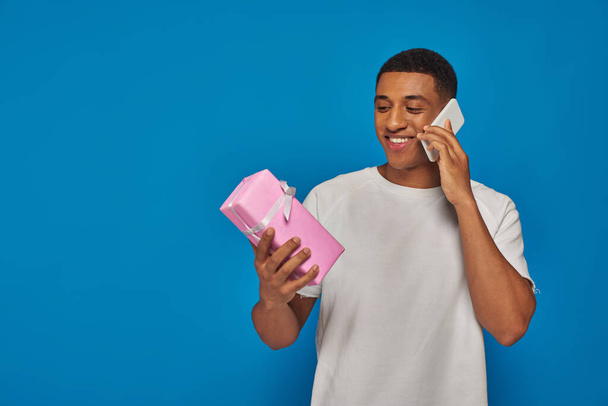 веселый африканский американец разговаривает на смартфоне и держит завернутый подарок на синем фоне - Фото, изображение