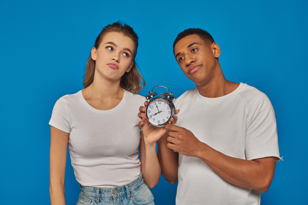 morning time, two displeased interracial couple holding retro alarm clock on blue backdrop - Photo, Image
