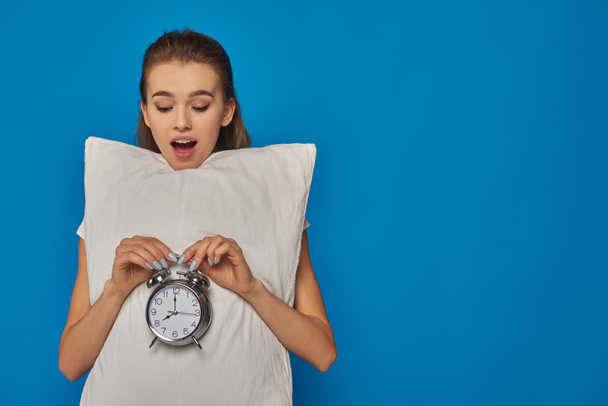 amazed young woman holding pillow and vintage alarm clock on blue background, early morning - Photo, Image
