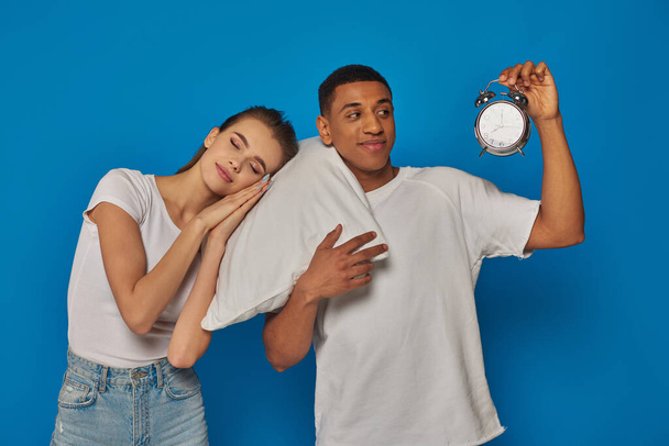 青い背景に目覚まし時計を持つ陽気なアフリカ系アメリカ人男性の近くの枕で眠っている女性 - 写真・画像