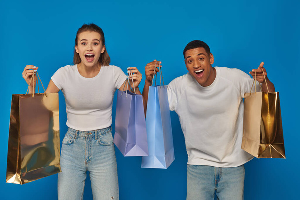 excitada pareja interracial sosteniendo bolsas de compras y mirando a la cámara en el fondo azul, consumismo - Foto, Imagen