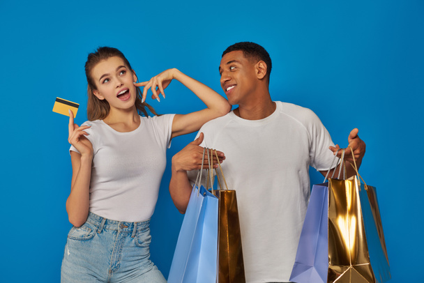ενθουσιασμένοι διαφυλετικό ζευγάρι κρατώντας τσάντες ψώνια και πιστωτική κάρτα σε μπλε φόντο, καταναλωτισμός - Φωτογραφία, εικόνα