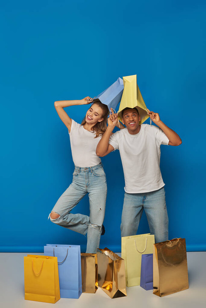 pareja multicultural positiva en atuendo casual sosteniendo bolsas de compras sobre fondo azul, alegría al por menor - Foto, Imagen