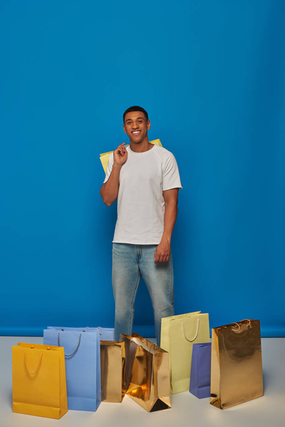 gioioso afro americano uomo in abbigliamento casual che tiene borse della spesa su sfondo blu, l'acquisto di furia - Foto, immagini