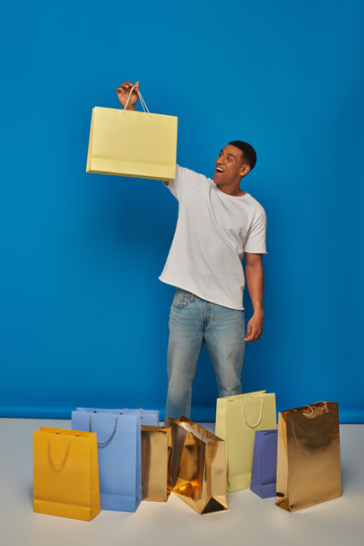 aufgeregter afrikanisch-amerikanischer Mann in lässiger Kleidung mit Einkaufstüten vor blauem Hintergrund, Kaufrausch - Foto, Bild