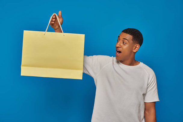 izgatott afro-amerikai férfi alkalmi öltözékben tartó bevásárló táska kék háttér, kiskereskedelmi öröm - Fotó, kép