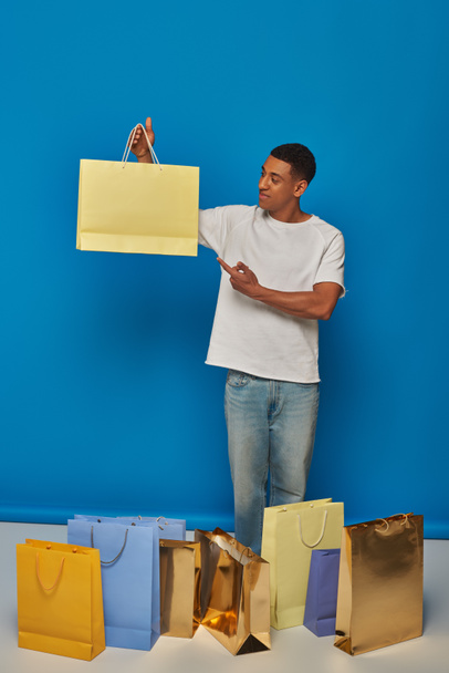 felice uomo africano americano in abbigliamento casual che punta alla borsa della spesa su sfondo blu, acquistare furia - Foto, immagini