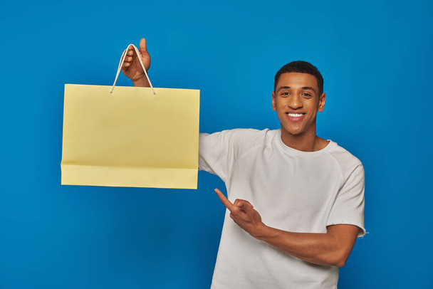 Radosny, afrykański Amerykanin w luźnym ubraniu, wskazujący na torbę na niebieskim tle, kupujący szaleństwo. - Zdjęcie, obraz