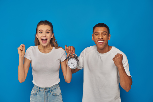 excited interracial couple with open mouth holding alarm clock on blue background, face expression - Photo, Image