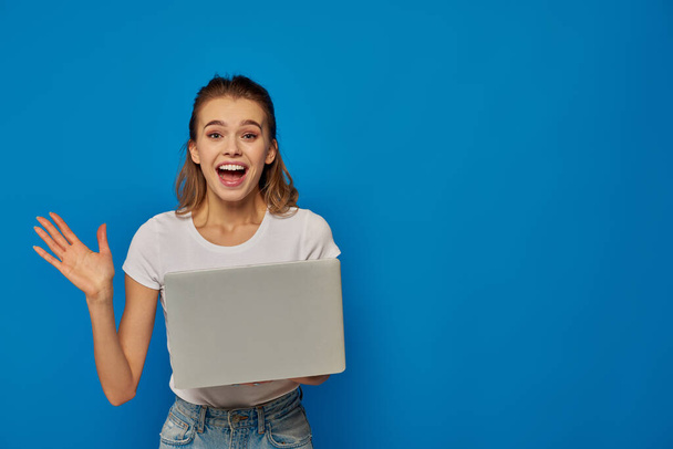 mujer excitada con el pelo rubio animando cerca del ordenador portátil sobre fondo azul, concepto de trabajo remoto - Foto, Imagen