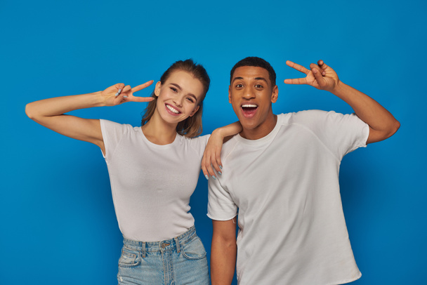 happy multicultural couple showing peace sign and looking at camera on blue background, positivity - Foto, immagini