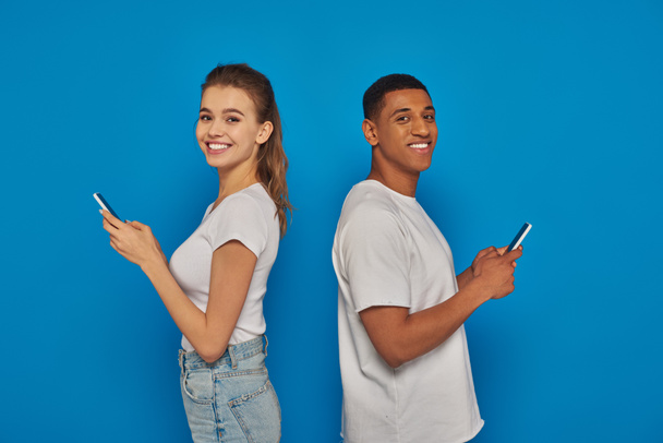 alegre pareja multicultural de pie espalda con espalda y el uso de teléfonos inteligentes sobre fondo azul - Foto, Imagen