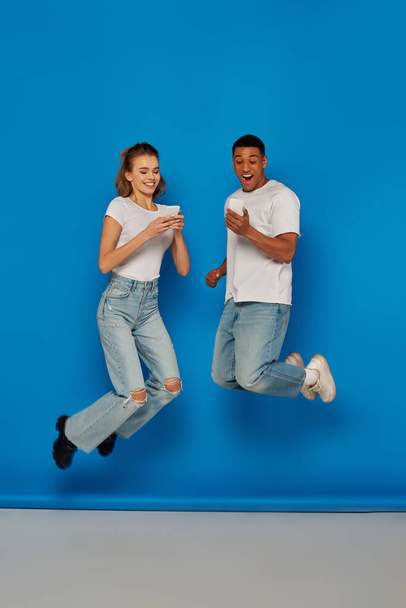 feliz casal multicultural usando smartphones e pulando em fundo azul, visão de comprimento total - Foto, Imagem