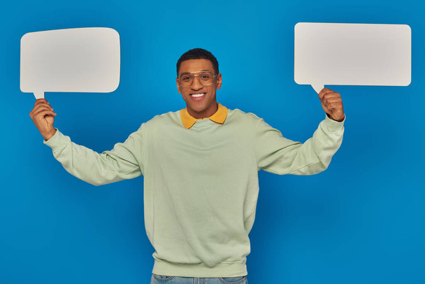 cheerful african american man in eyeglasses and smart casual clothes holding blank speech bubbles - Photo, Image