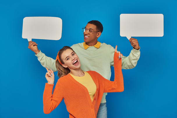 cheerful diverse couple in smart casual clothes holding blank speech bubbles on blue backdrop - Fotoğraf, Görsel