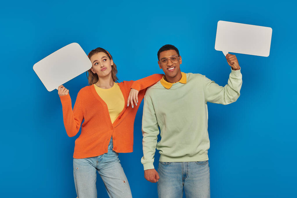 cheerful interracial couple in smart casual clothes holding blank speech bubbles on blue backdrop - Photo, Image