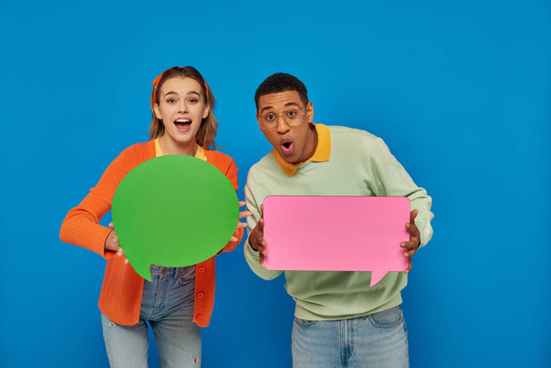 excited interracial couple in smart casual clothes holding blank speech bubbles on blue backdrop - Photo, Image