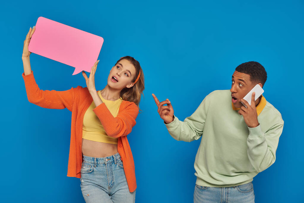 amazed african american man in glasses pointing at woman with speech buttle on blue background - Photo, Image
