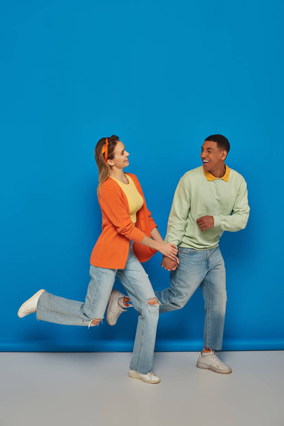 ευτυχισμένο πολυπολιτισμικό ζευγάρι σε casual ενδυμασία κρατώντας τα χέρια και τρέχει σε μπλε φόντο, χαμόγελο - Φωτογραφία, εικόνα