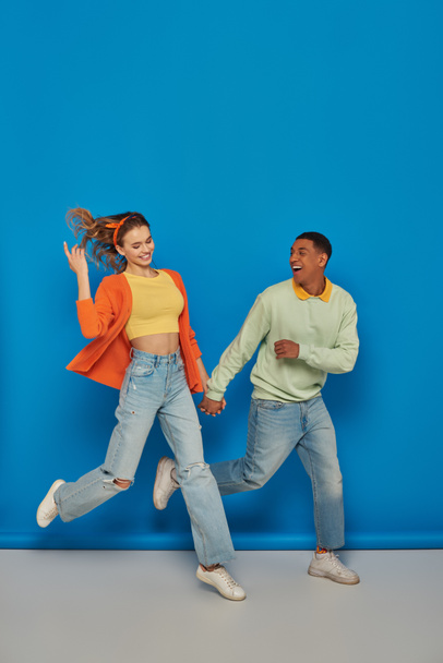alegre pareja multicultural en traje casual cogido de la mano y corriendo sobre fondo azul, sonrisa - Foto, Imagen