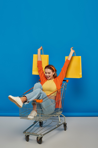 excited young woman sitting in cart and holding shopping bags on blue background, buying spree - Photo, Image