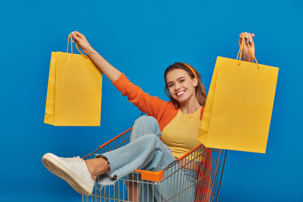 positieve jonge vrouw zitten in winkelwagen en het houden van boodschappentassen op blauwe achtergrond, kopen spree - Foto, afbeelding