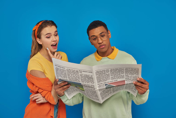 青い背景の眼鏡で落ち着いたアフリカ系アメリカ人の男性の近くで新聞を読んでいる驚いた女性 - 写真・画像