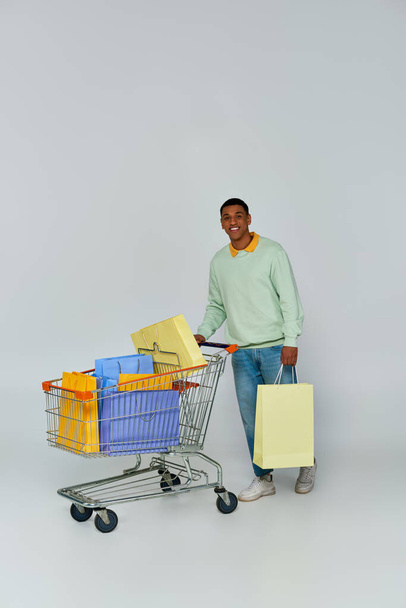 hombre afroamericano emocionado de pie con carro y bolsas de compras en el fondo gris, el consumismo - Foto, Imagen