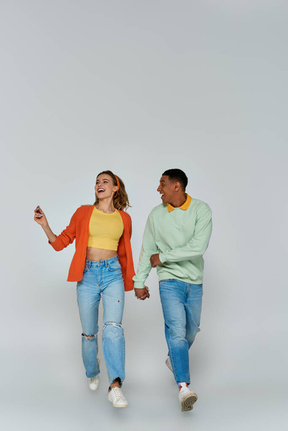 glückliches interrassisches Paar in lässiger Kleidung, Händchen haltend und vor grauem Hintergrund zusammen laufend - Foto, Bild