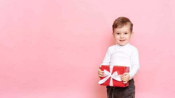 Подарунок у руки хлопчику. Щасливий хлопчик з подарунковою коробкою в руках ізольований рожевий фон
. - Фото, зображення