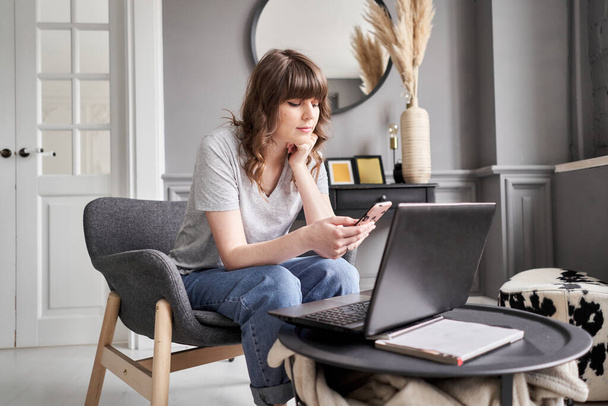 Schöne fröhliche Mädchen mit schwarzem Laptop, während sie auf dem Sofa im Wohnzimmer zu Hause sitzt. Junge Gelegenheitsfrau mit Laptop für Kommunikation und Arbeit. S - Foto, Bild