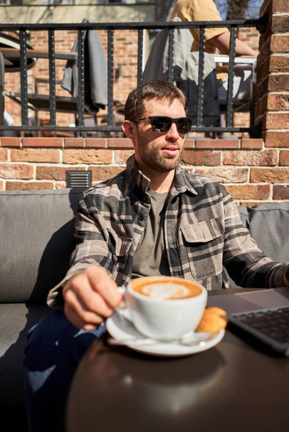 Чоловік п'є каву, тримає в руці чашку гарячого арахісового латте. Стіл у коворкінгу кав'ярні. Чашка кави на веранді в сонячний день. Кавова перерва в кафе. Крупним планом - Фото, зображення