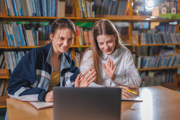Two teen girls taking online class instruction using a laptop while sitting in a school library. Education, teaching, and technology concepts. - Valokuva, kuva
