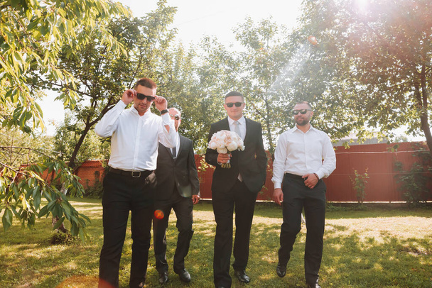 The groom in a black suit and glasses and his stylish friends wearing white shirts and black pants and glasses are standing in the backyard in the garden. The groom is holding a bouquet. - Fotó, kép