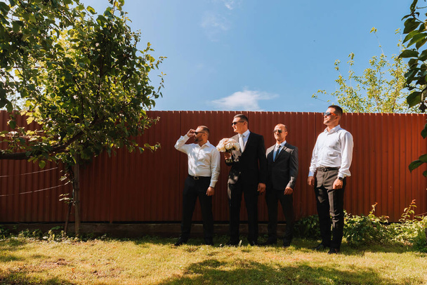 The groom in a black suit and glasses and his stylish friends wearing white shirts and black pants and glasses are standing in the backyard in the garden. The groom is holding a bouquet. - Foto, imagen
