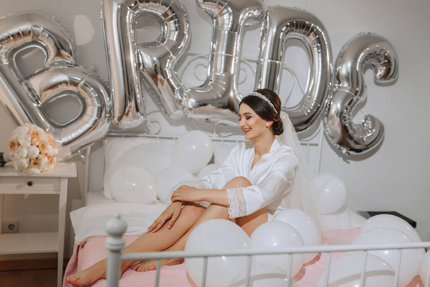 A beautiful brunette bride in a white robe, a veil and a tiara on her head, sits on a bed. Inscription bride. Happy young girl under the glow of sunlight. wedding concept - Fotó, kép