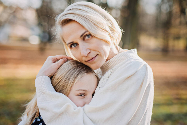 Blond moeder en tienerdochter knuffelen op herfstpark op een zonnige dag. Moederlijke liefde en steun voor tieners. - Foto, afbeelding