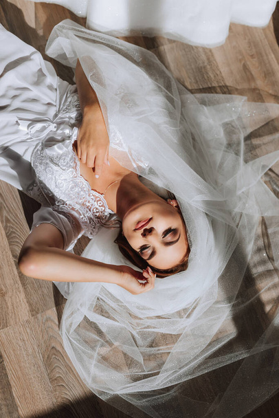 A beautiful young bride in a white robe poses for a photographer, lying on the floor, on a veil. Wedding photography, close-up portrait, chic hairstyle. - Foto, imagen