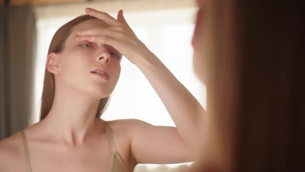 Anti-aging cosmetica. Vrouw brengt anti-rimpel crème op het gebied in de buurt van de ogen Kaukasische jonge dame kijkt in de spiegel Schoonheidsroutine Gezichtsverzorging - Video
