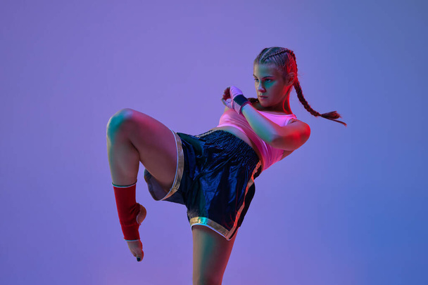 Leg kick. Sportive teen girl, mma fighter athlete in motion, training, fighting against purple background in neon lights. Concept of mixed martial arts, sport, hobby, competition, strength, ad - Foto, Imagen