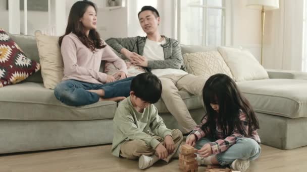 δύο παιδιά από την Ασία κάθονται στο πάτωμα παίζοντας ενώ οι γονείς συνομιλούν στον καναπέ στο παρασκήνιο στο σπίτι - Πλάνα, βίντεο