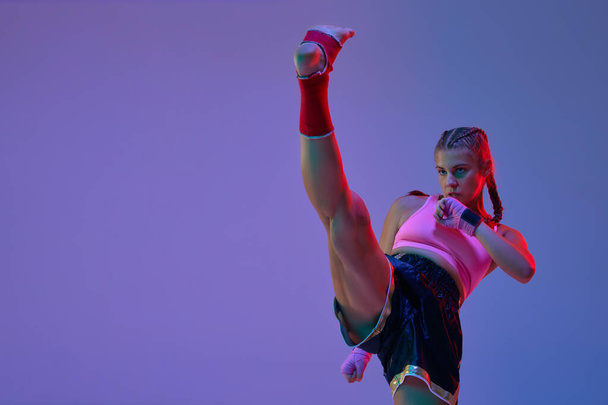 Leg kick. MMA fighter, teen girl training, fighting against purple studio background in neon lights. Concept of mixed martial arts, sport, hobby, competition, athleticism, strength, ad - Photo, image